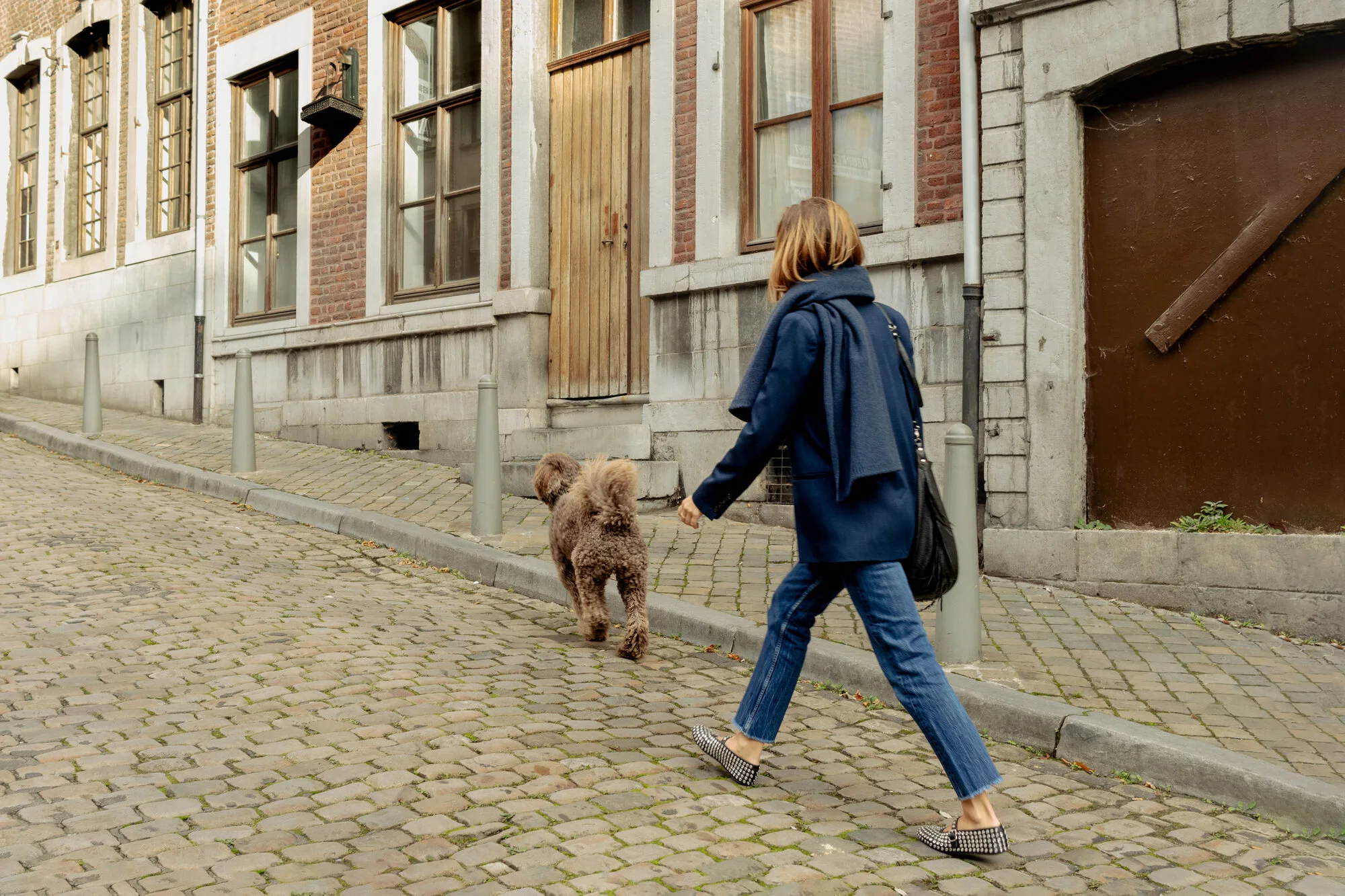 Une femme marche avec son chien dans une rue pavée, portant un manteau bleu et une étole bleu marine