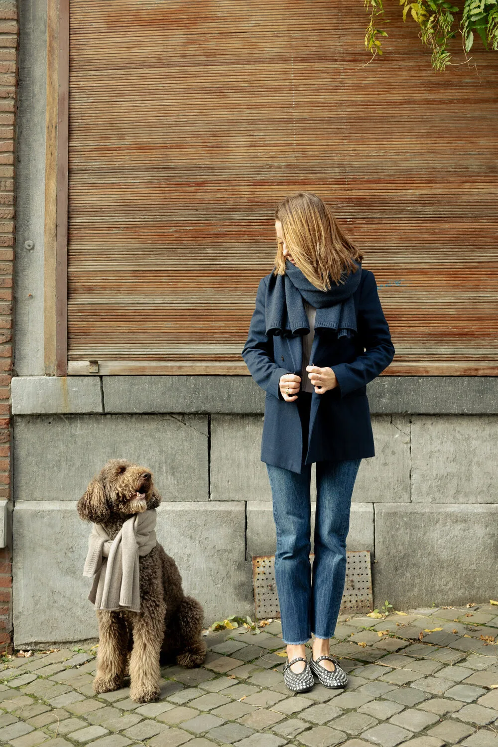 Une femme regarde son chien assis, habillé d'une écharpe beige, dans une rue pavée.